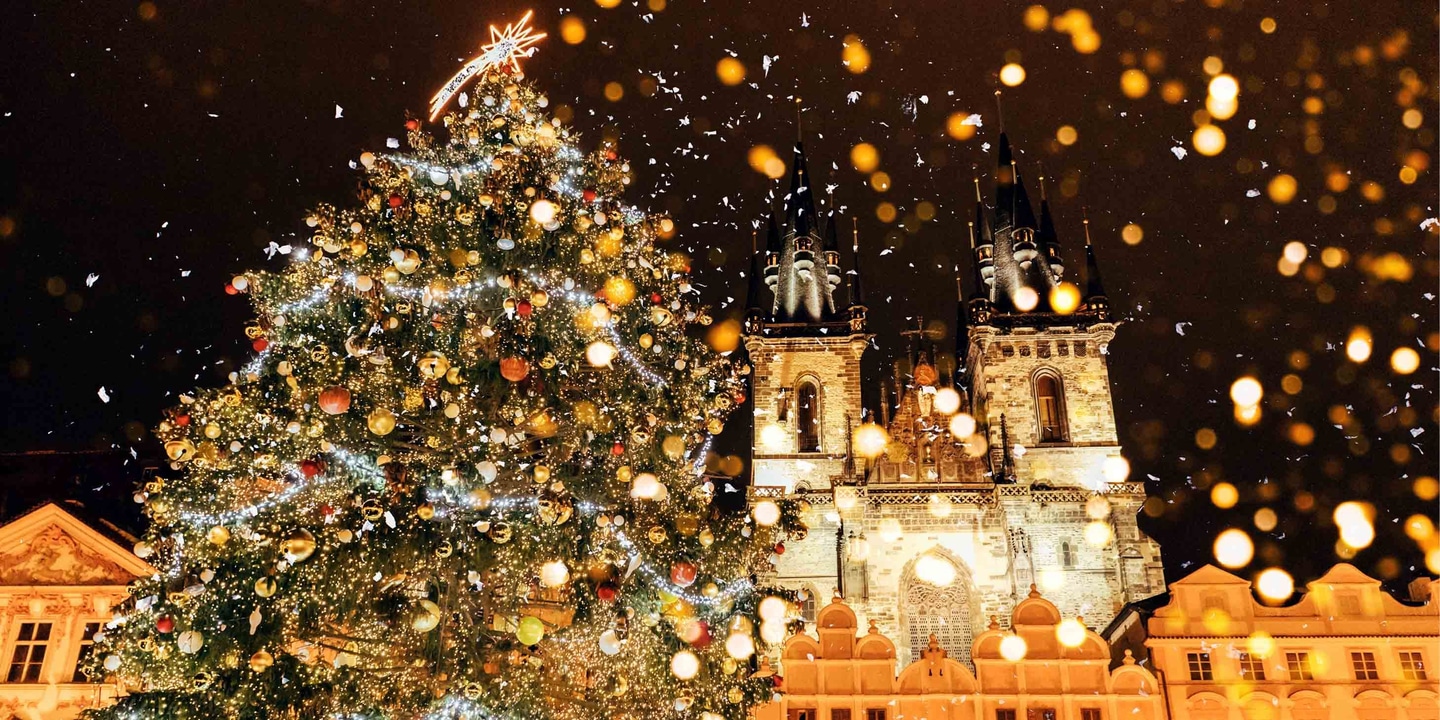 A christmas tree covered with lights, baubles and decorations in front of an illuminated building in Prague.