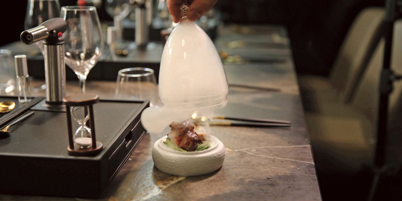 The waiter lifts a smoke filled glass above a dish, showcasing the prepared meal in a focused manner.