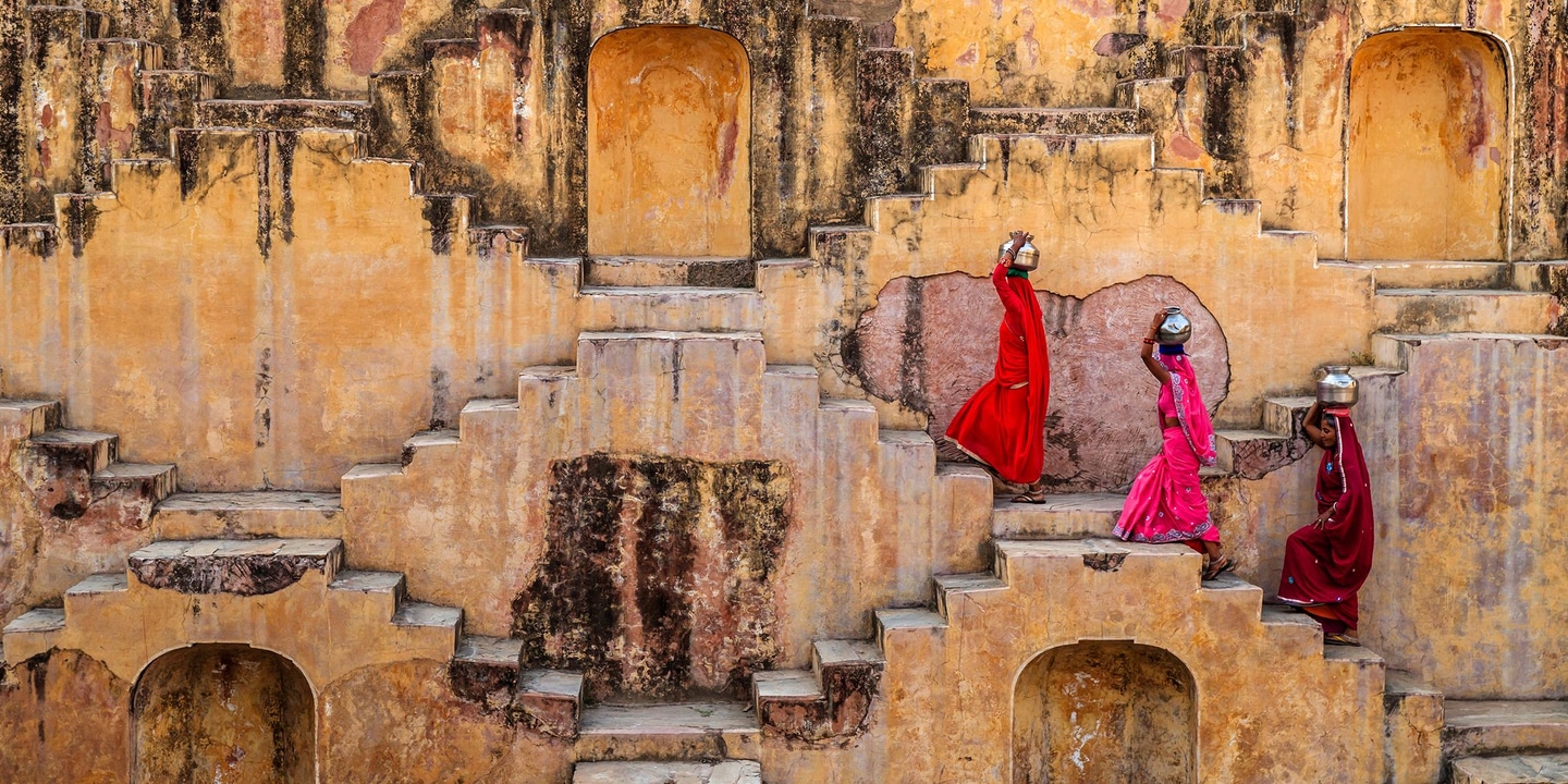 Three women in vibrant red and pink saris walking along the geometric steps and niches of an old sandstone stepwell in Jaipur, India, creating a striking pattern of colour against the weathered walls.